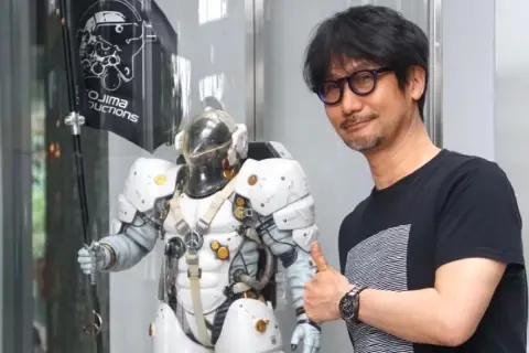 Hideo Kojima z kciukiem w górę obok postaci z gry, z logo Kojima Productions w tle.