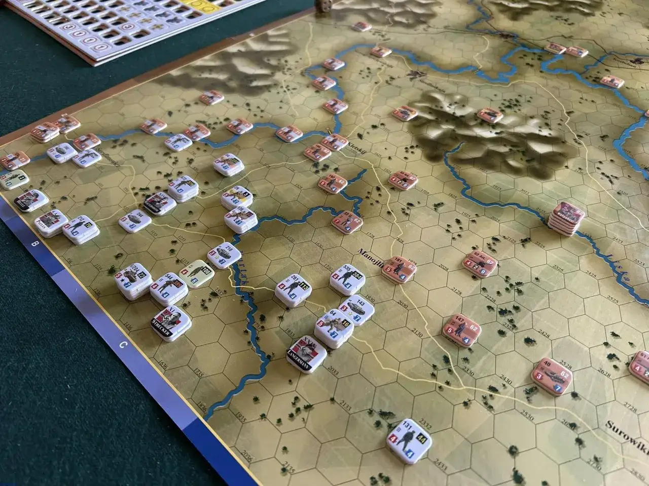 Plansza gry strategicznej z żetonami wojsk, rzeką i wzgórzami. Widać fragmenty mapy, które przywołują klimat old world.