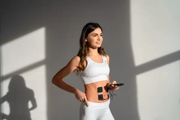Elin w białym sportowym topie i legginsach, z elektrodami na brzuchu, trzyma smartfon.